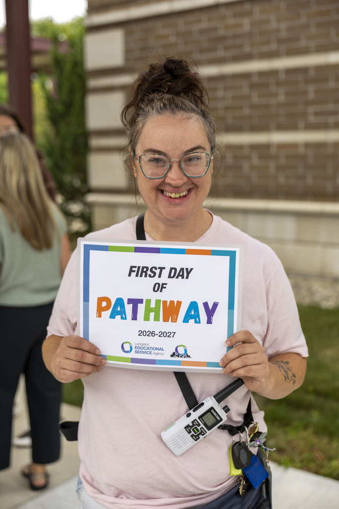 A smiling woman with glasses and a messy bun holds a "First Day of Pathway 2026–2027" sign.