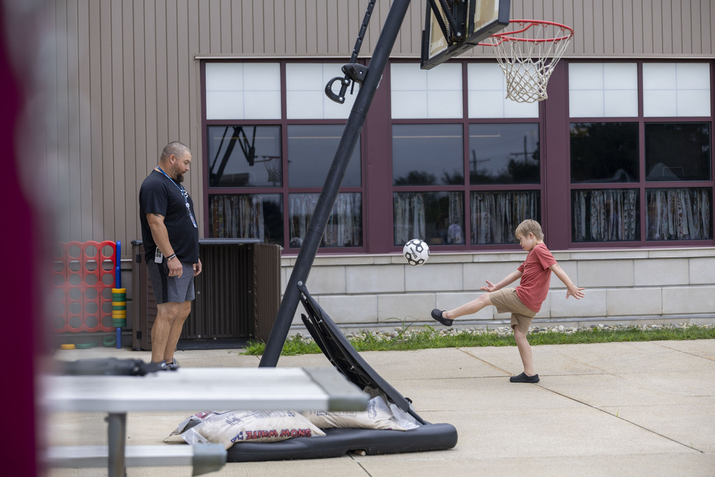 A young boy kicks a soccer ball high in the air near a portable basketball hoop while an instructor observes.