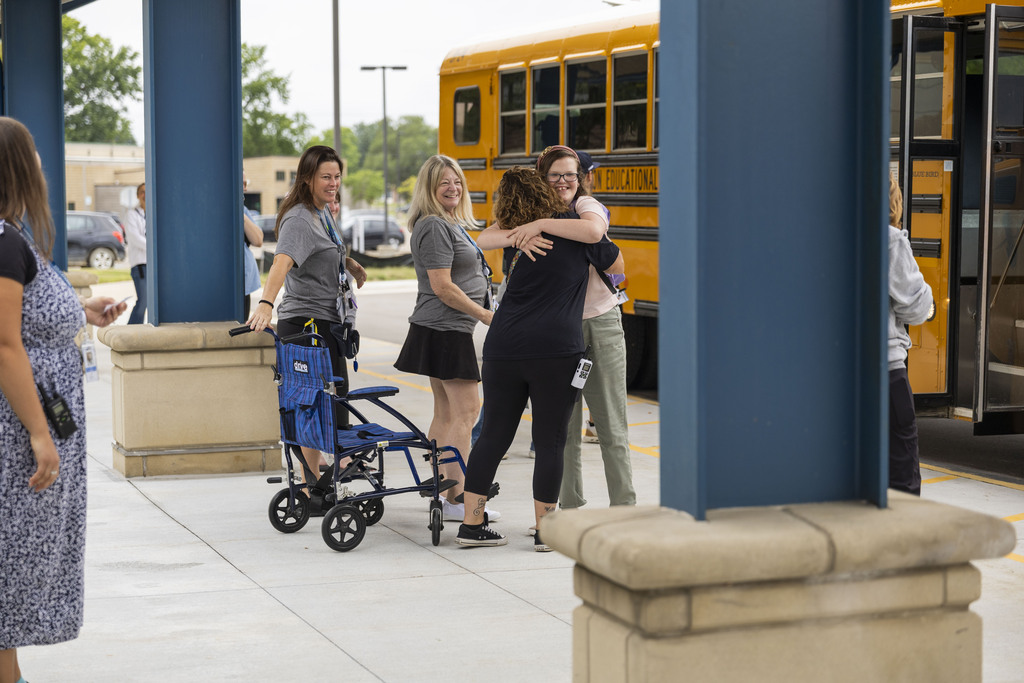 School staff warmly welcome a student with a hug as she steps off a yellow school bus.