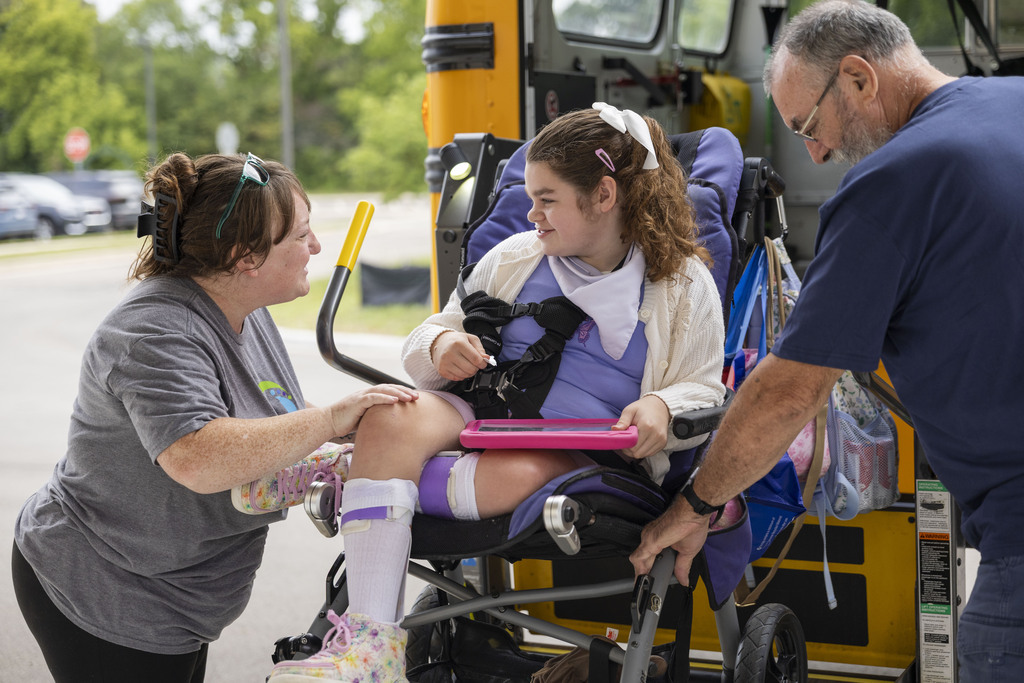 School staff assist a young girl in a specialized wheelchair as she exits a bus lift.