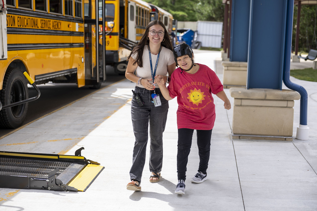 A female staff member holds hands with a smiling student wearing a helmet as they walk along the bus loading area.