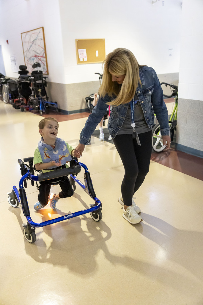 A female staff member holds hands with a smiling young boy using a blue gait trainer down a hallway.