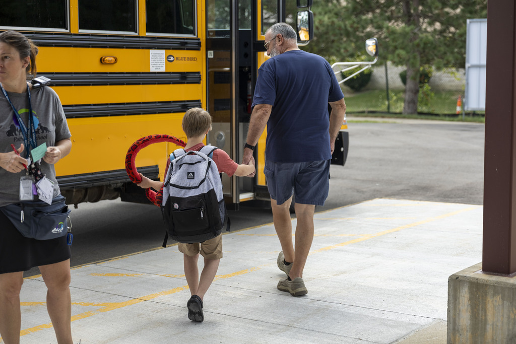 A young boy holding a red stuffed snake walks hand-in-hand with an adult towards a yellow school bus.
