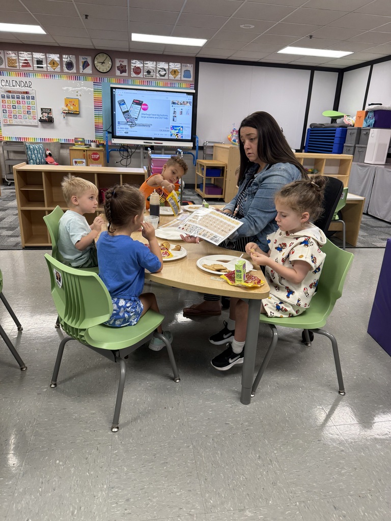 GSRP students sit around a classroom table eating breakfast while a teacher holds and uses a mobile COR communication board with the children.
