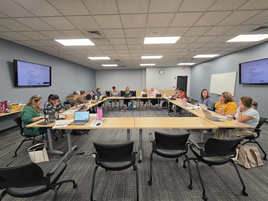 Participants gathered around an O-shaped table arrangement inside a conference room with wall-mounted screens, whiteboards, and overhead recessed lighting, actively engaged in a group meeting or workshop.
