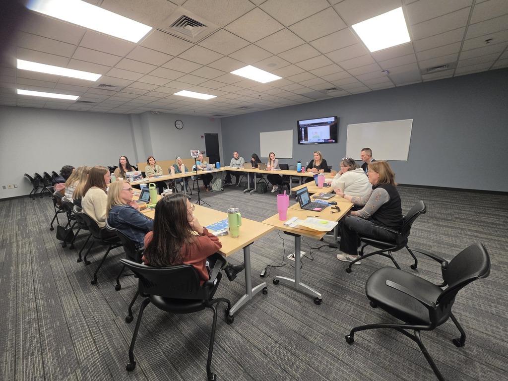 A group of people seated around an O-shaped table setup in a modern conference room, working on laptops during a meeting.