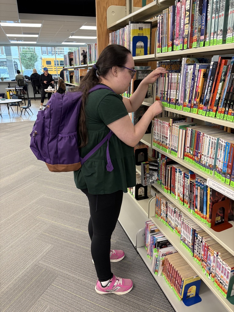 A student with a purple backpack browses books on a shelf in a library.