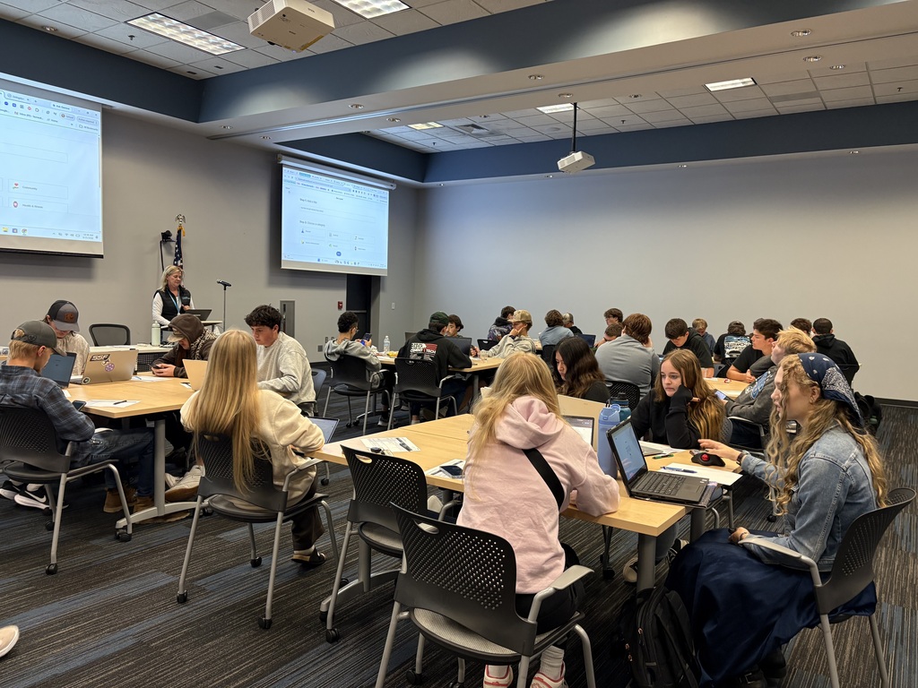 A classroom filled with students working in small groups at tables, with laptops and papers in front of them. An instructor stands near a lectern, while two projectors display computer screens on the front and side walls.
