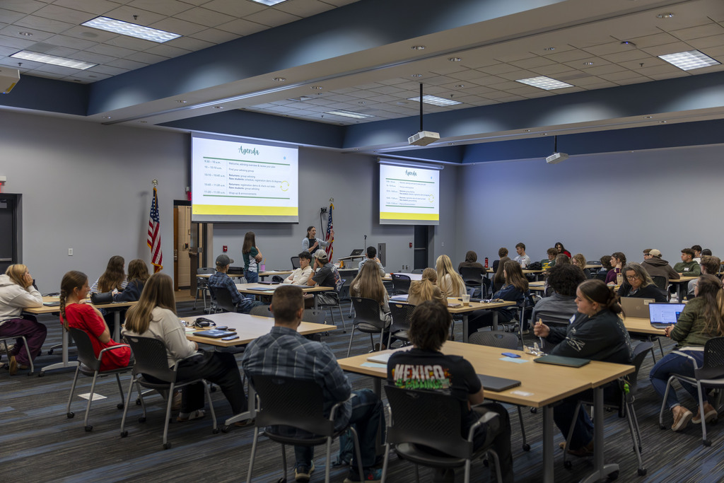A classroom full of students seated at tables during a presentation, with an instructor at the front, American flags, and two projection screens displaying an agenda.