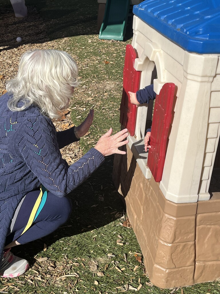 High-angle view of a woman with short, wavy blonde hair wearing a blue knit sweater sitting outside on the grass, reaching up with an open hand toward a child inside a plastic playhouse. The child’s hands peek out through the playhouse’s red window shutters, playfully interacting and playing peek-a-boo with the woman.