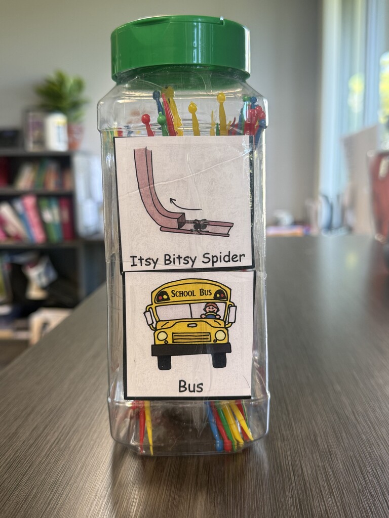 A clear plastic container filled with colorful plastic picks, repurposed as an educational activity tool. Two visual picture communication cards are taped to the front: the top card shows an illustration labeled "Itsy Bitsy Spider," and the bottom card shows a yellow school bus labeled "Bus."
