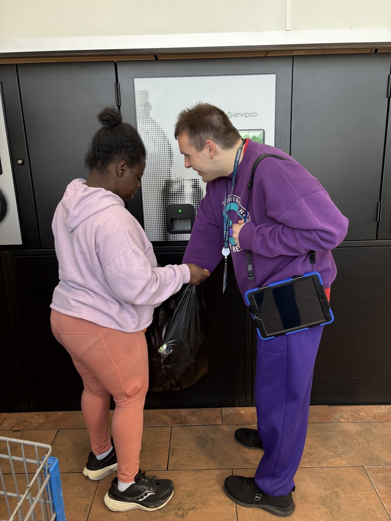 Two people standing near a bottle return machine, holding a bag of bottles and cans to deposit.