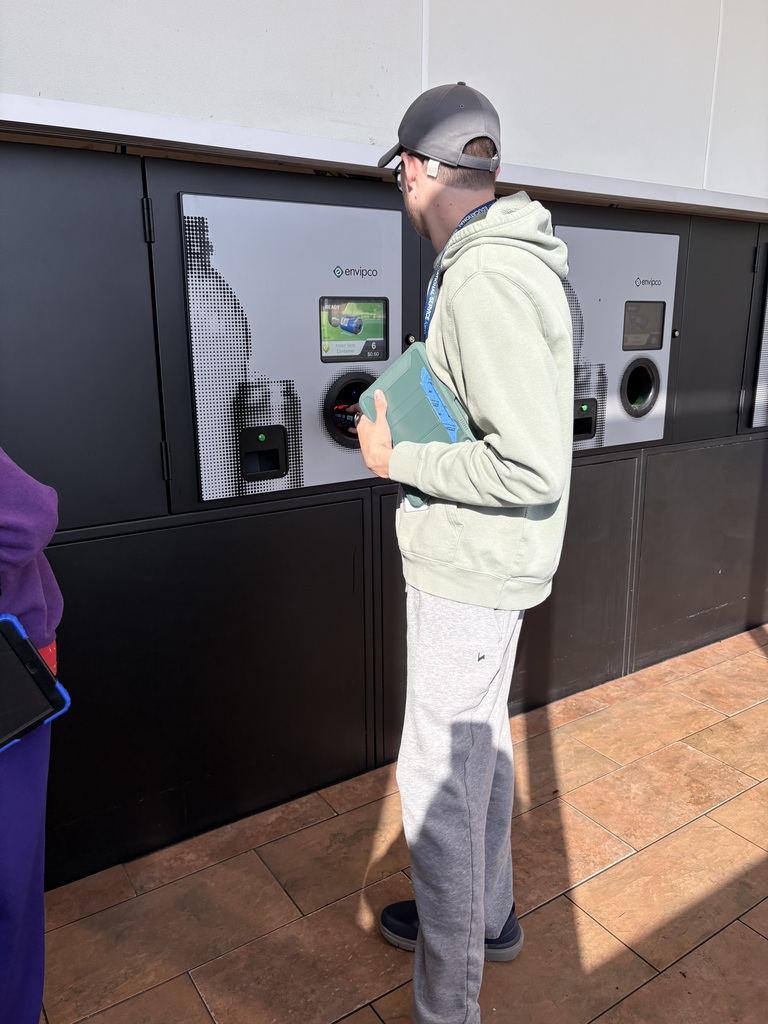 A person in a baseball cap and light green hoodie inserting a bottle into a bottle return machine.