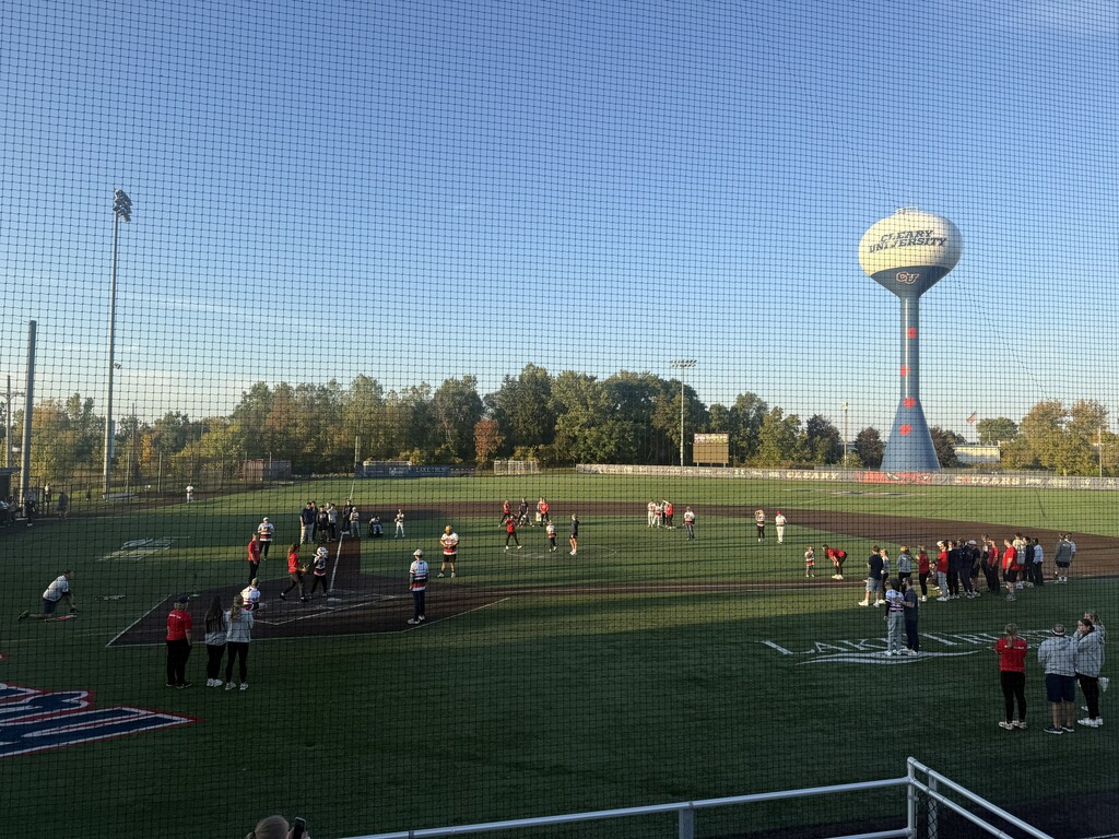 Wide shot taken through a protective netting of a baseball or softball field where teams and spectators are gathered on the turf under a clear blue sky, with a tall Cleary University water tower visible in the background.