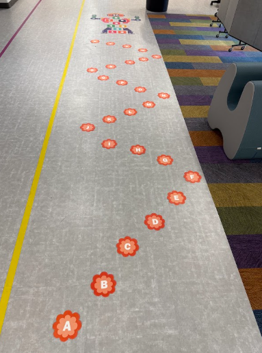 An alphabetical floor path made of orange flower decals winding along a hallway toward a robot hopscotch game.