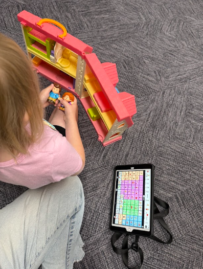 Top-down view of a child sitting on a patterned carpet floor playing with toy figures and a pink dollhouse, with an AAC speech-generating device on a tablet lying nearby.
