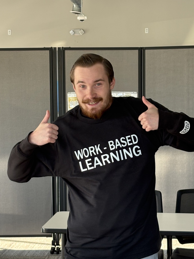 A young man smiling and giving two thumbs up while wearing a black long-sleeve shirt that reads "WORK - BASED LEARNING".