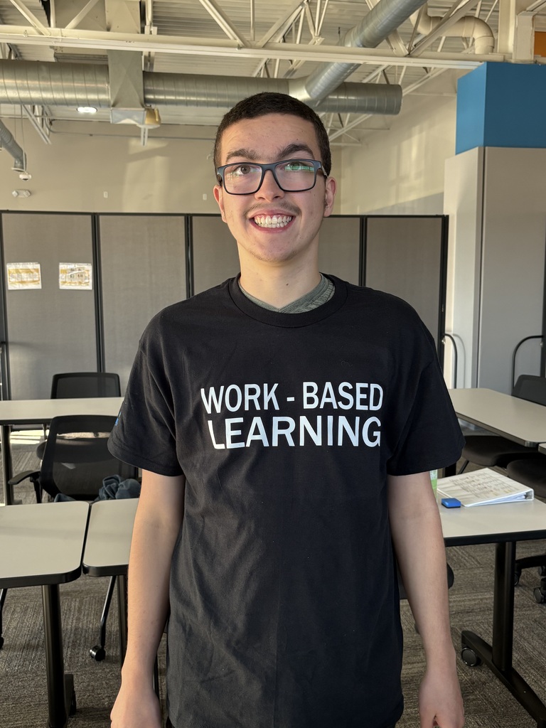A smiling young man wearing glasses and a black t-shirt that reads "WORK - BASED LEARNING".