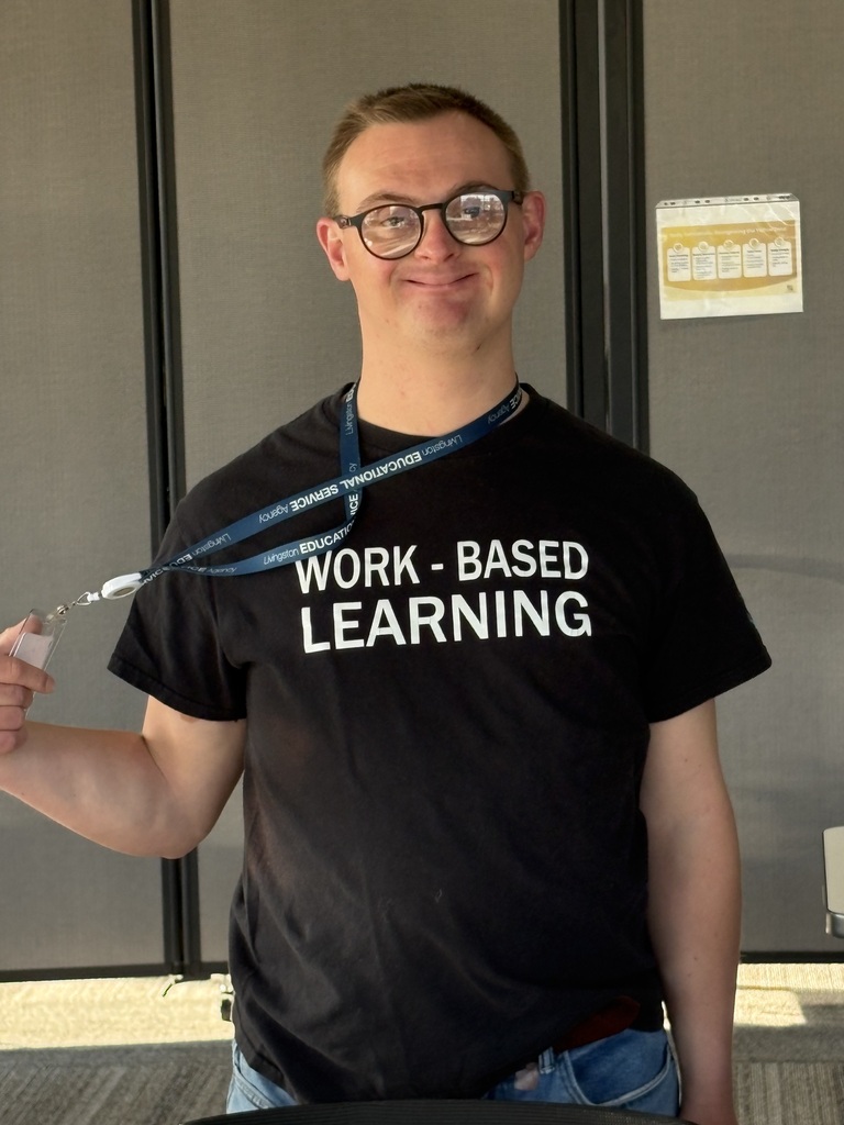 A smiling young man wearing glasses and a black t-shirt that reads "WORK - BASED LEARNING".