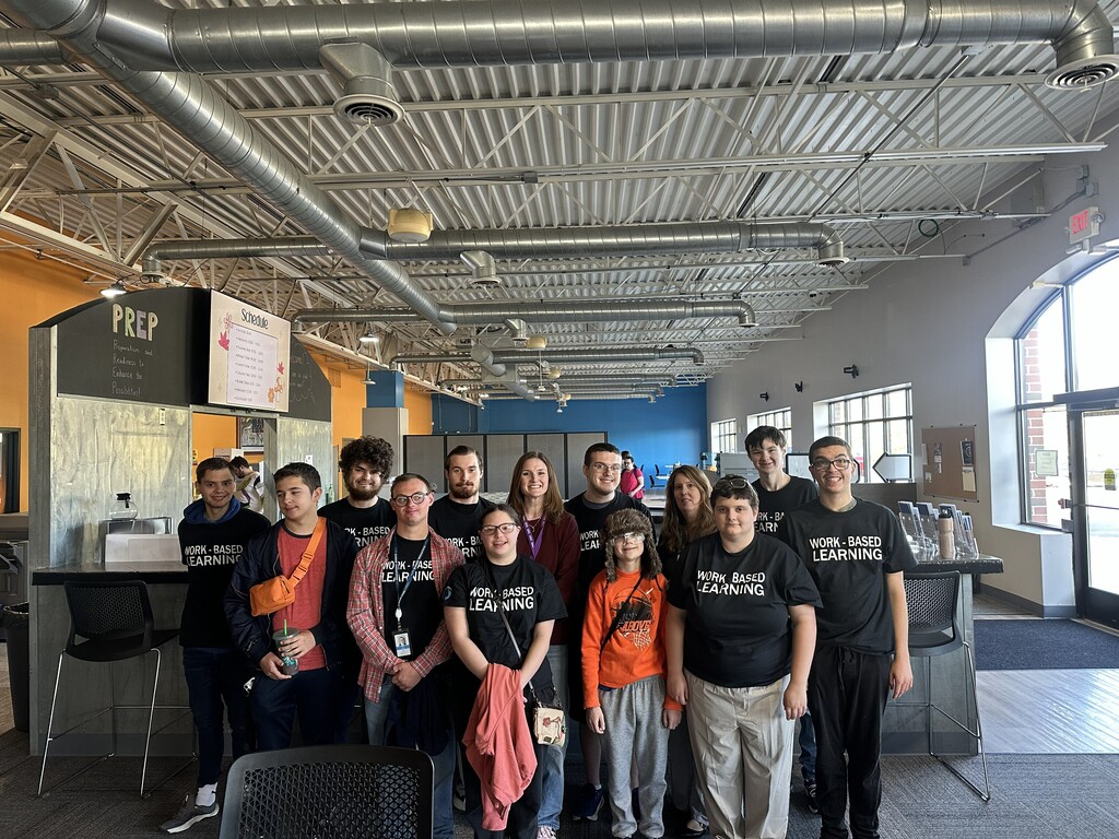 A group of students and instructors posing together in a building lobby, with the students wearing matching black "WORK - BASED LEARNING" shirts.