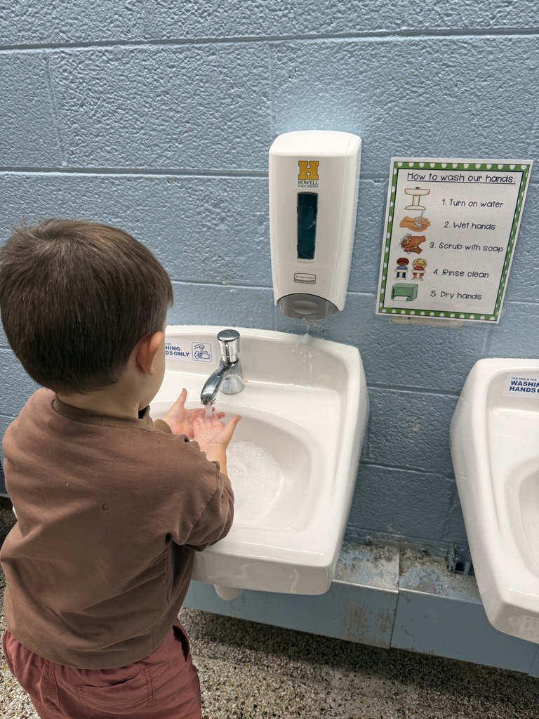 A young child washes his hands at a classroom sink. A visual handwashing guide posted beside the sink shows five illustrated steps to support independence.