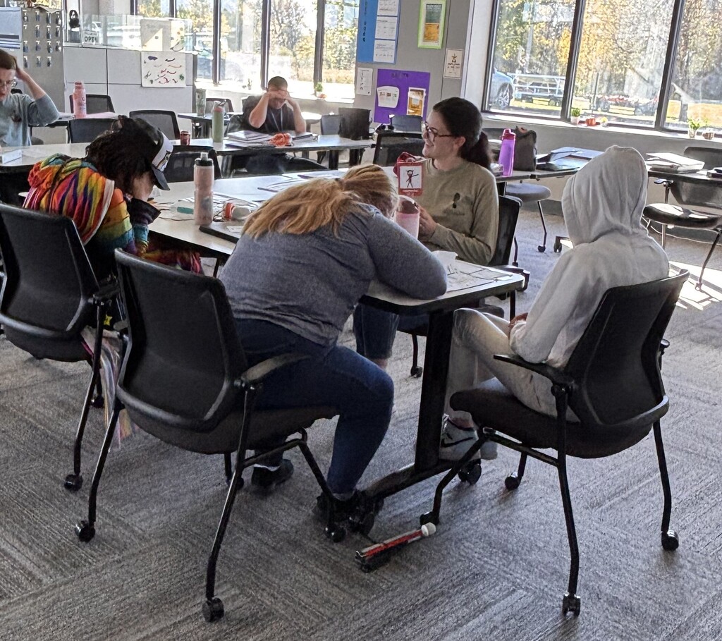 A small group of students leaning over desks to collaborate on an assignment with a instructor looking on.