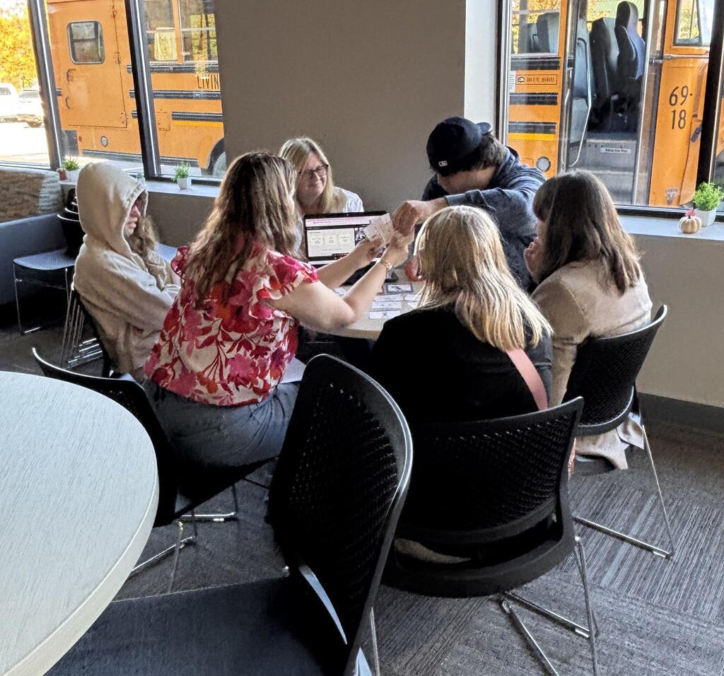 A small group of students and staff sitting around a table with a laptop, sharing learning materials and cards.