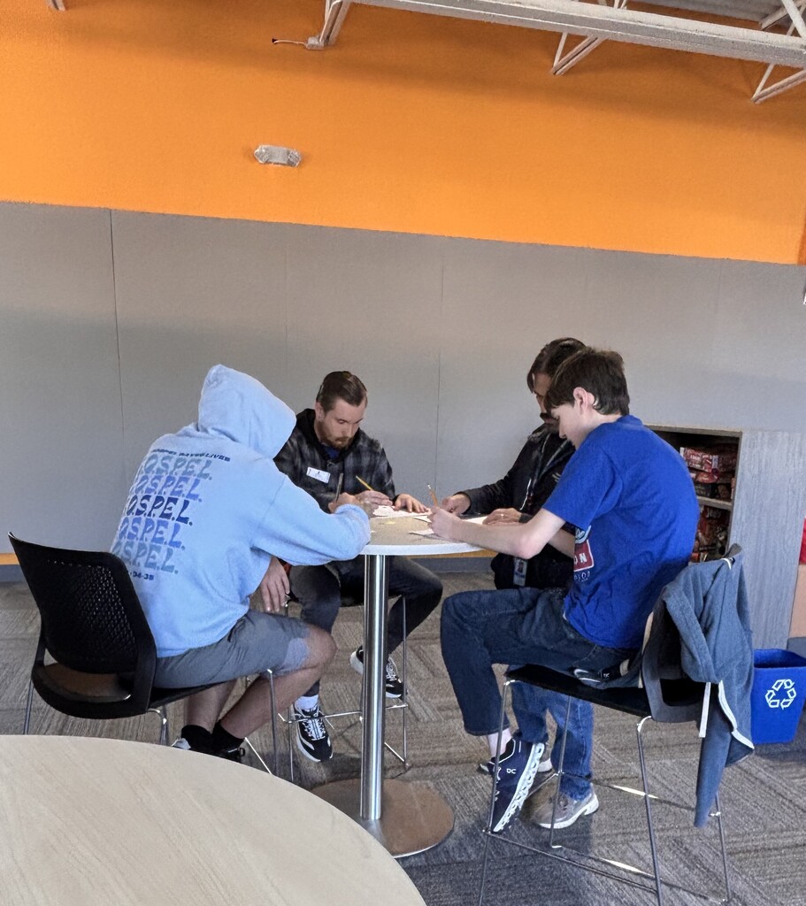 A group of students and a staff member sitting around a small high-top table working together with pencils and papers.