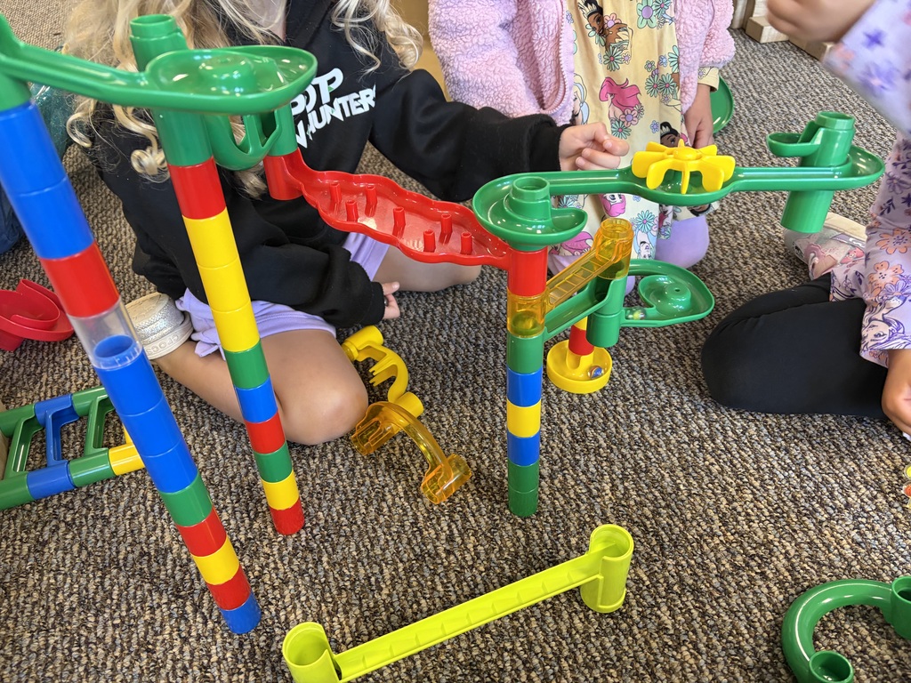 Preschool children work together to build a colorful marble run using connecting tubes and tracks, exploring problem-solving, building, and cause-and-effect through hands-on play.