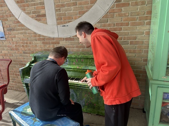 A student playing a painted outdoor piano while another student in a red hoodie leans over to watch and smile.
