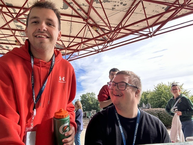 Two young men smiling together outdoors under a covered pavilion at a park.