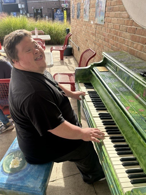 A man smiling joyfully while playing a brightly painted green outdoor piano sitting on a decorated blue bench.