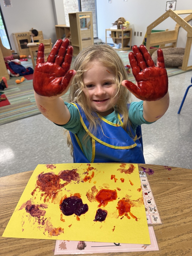A smiling child holds up both hands covered in red paint, with a colorful handprint painting on yellow paper in front of them.