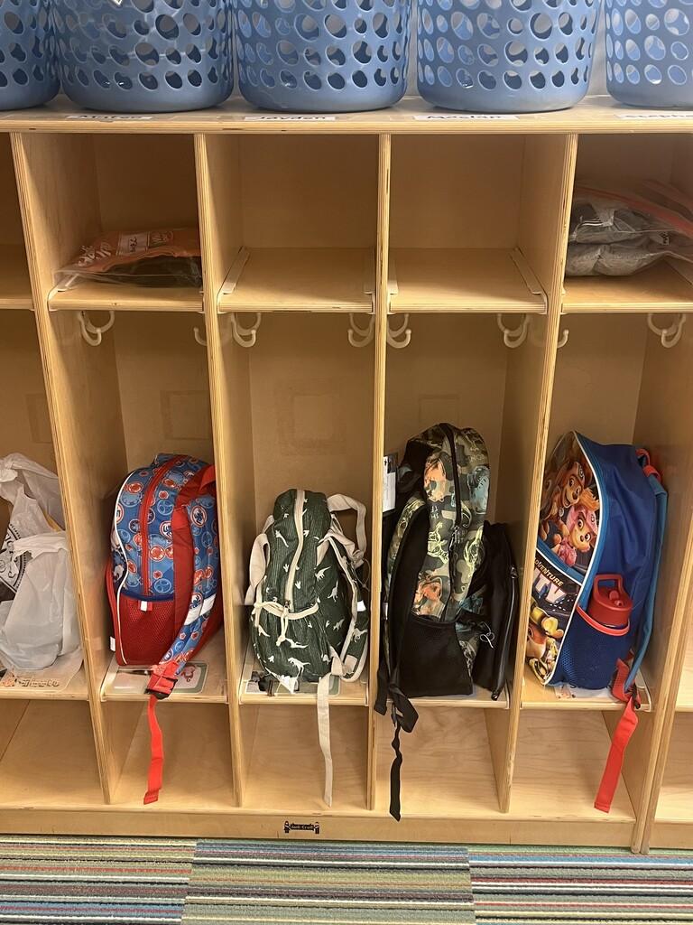 A set of light wood classroom cubbies lined up against a wall. The top shelf holds several blue perforated plastic storage baskets. In the cubby compartments below, four kids' backpacks sit on the bottom shelves—including a red and blue Spider-Man bag, a dark green backpack with white dinosaur silhouettes, a camouflage print backpack, and a blue Paw Patrol backpack—along with plastic bags stored on the top shelves.