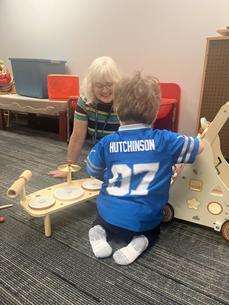 A child wearing a blue Detroit Lions football jersey with "HUTCHINSON" and number 97 on the back sits on a gray carpeted floor, playing with a small wooden toy drum set. A smiling adult with blonde hair and glasses kneels beside the child near a wooden activity walker and colorful play equipment. 