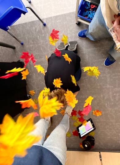 High-angle view of a child kneeling head-down on a grey tiled floor, with vibrant red, orange, and yellow fake leaves showering down and landing on their back and head. Adults stand around the child, and a tablet displaying a communication grid lies on the floor nearby amid scattered leaves.