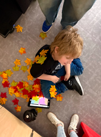 Top-down view of a young boy with light hair sitting cross-legged on a grey tiled floor, wearing a black t-shirt and blue jeans. Fake red, orange, and yellow autumn leaves are resting on his shoulder, on his leg, and piled beside him near a tablet with a colorful communication grid displayed.