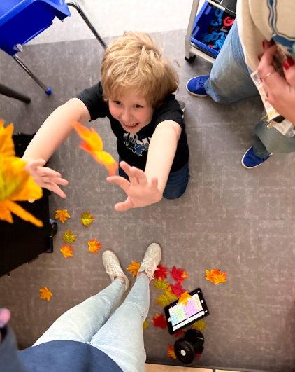 Top-down view of a joyful blonde-haired boy looking up and reaching out to catch fake orange and yellow autumn leaves falling around him. Colorful artificial leaves are scattered on the grey tile floor near an adult's feet and a tablet displaying a communication app.