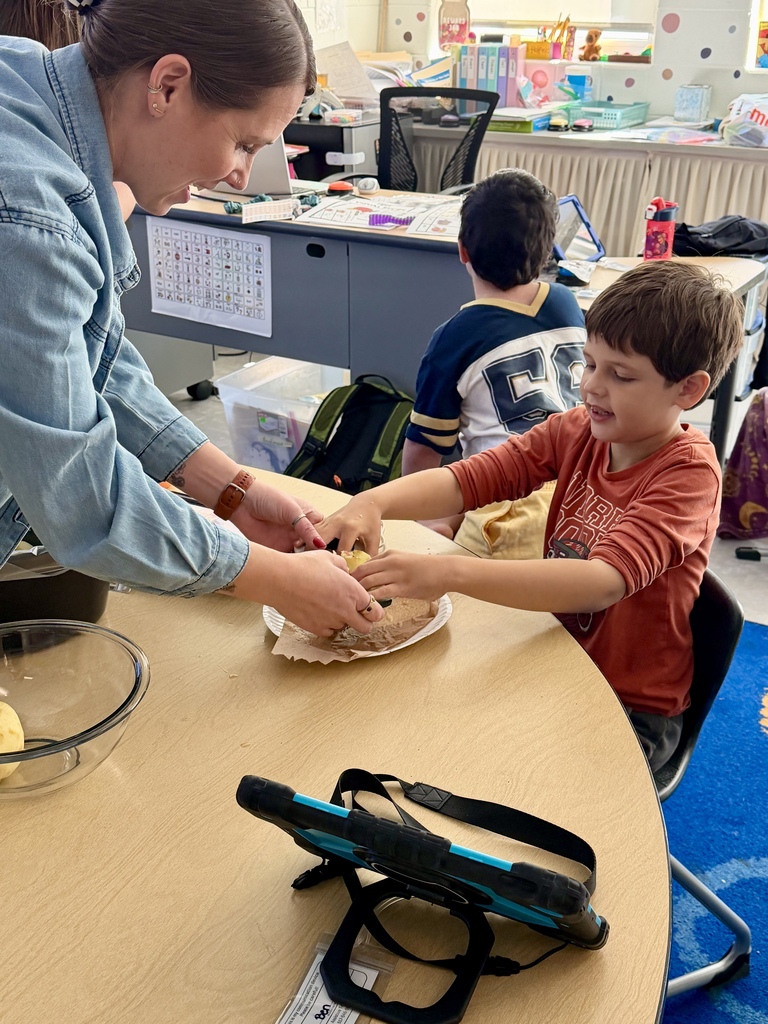 An OT in a denim jacket smiles as she helps a young boy sitting at a classroom table slice apples, with an AAC tablet resting in the foreground.