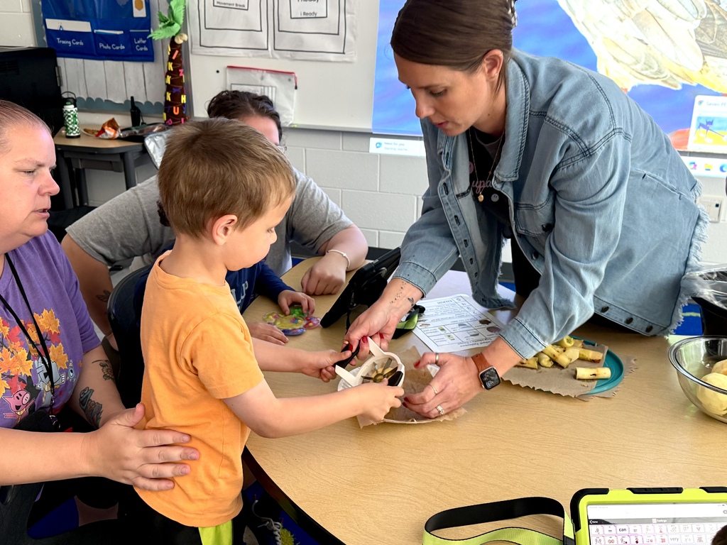 Side profile view of a young boy in an orange shirt standing at a classroom table, holding an apple slicer with assistance from an OT wearing a denim jacket. An adult in a purple shirt sits beside him for support, while sliced apple pieces, a recipe visual schedule, and an AAC tablet rest on the table around them.