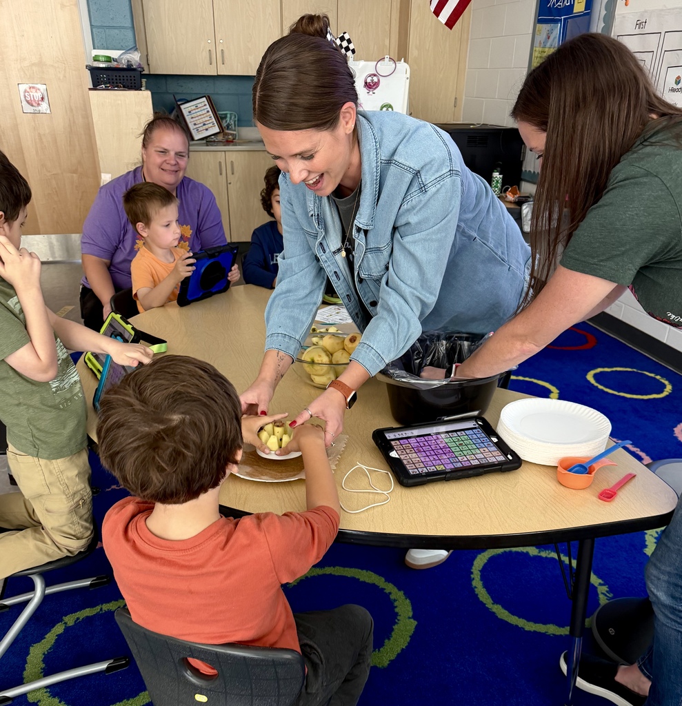 An OT in a denim jacket smiling as she helps a young boy push down on an apple slicer during a classroom activity, with an AAC tablet on the table nearby.