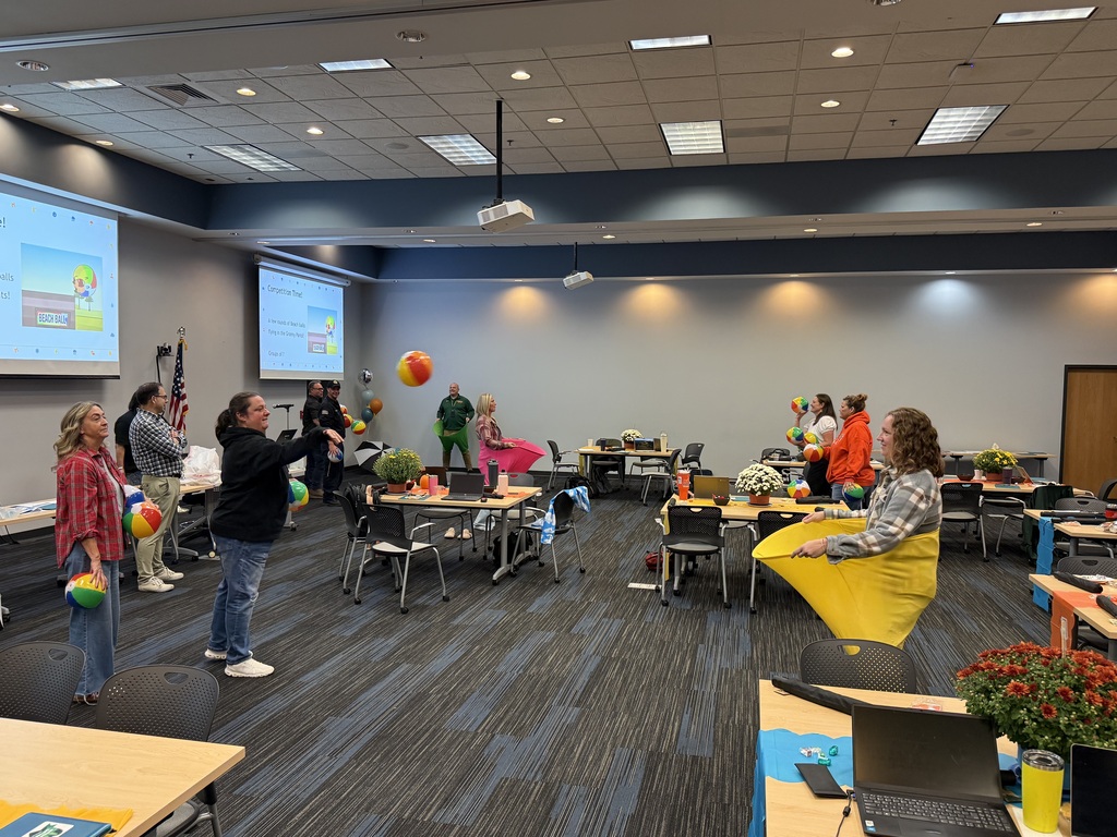  Adults participate in a group activity in a conference room, tossing colorful balls to one another while standing or sitting in large yellow fabric buckets. Tables, chairs, laptops, and presentation screens are arranged throughout the room.