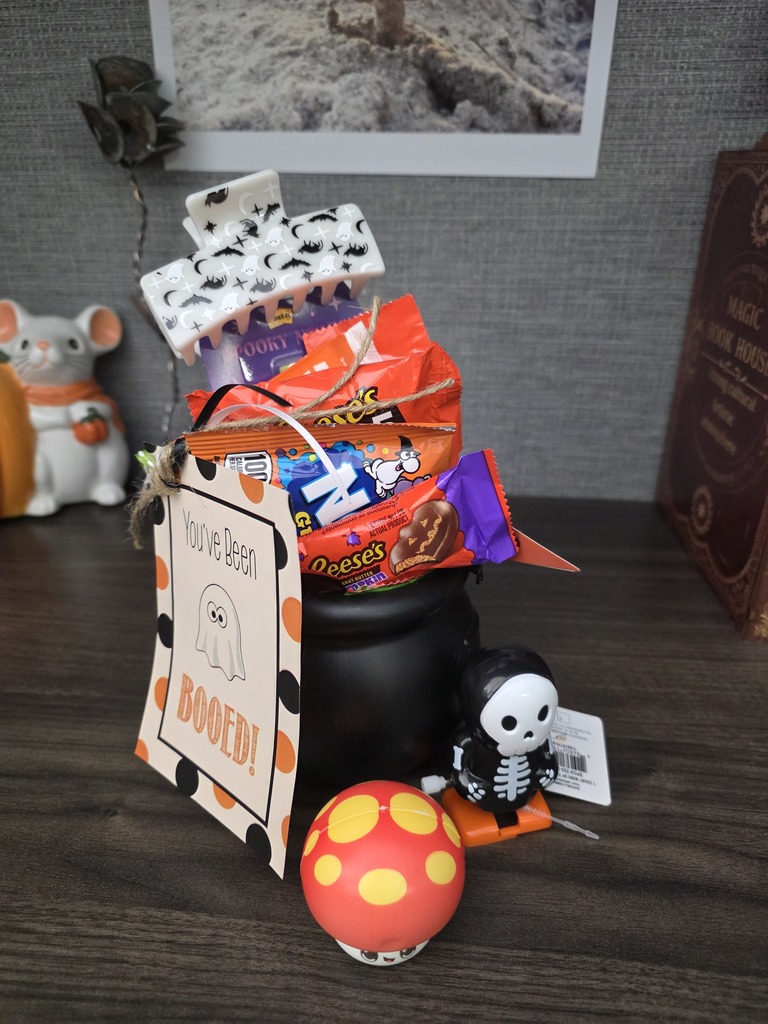 A small black plastic cauldron filled with candy, a hair clip, and other goodies with a tag reading "You've been Booed!" resting on a desk.