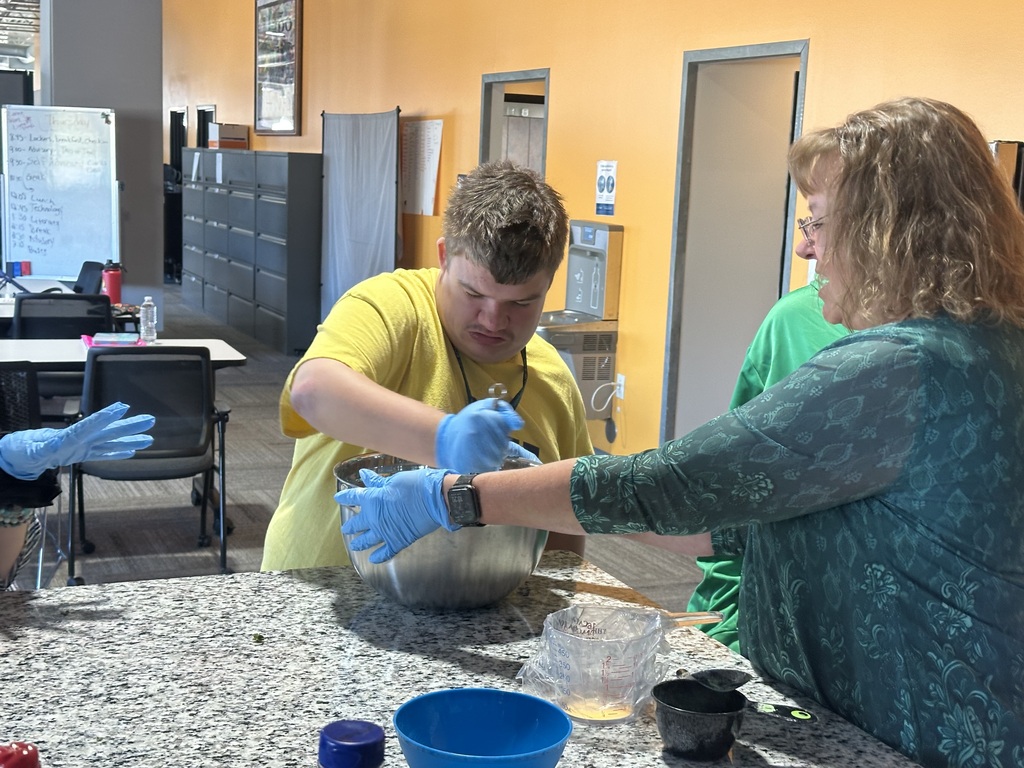 A young man with blue gloves mixes ingredients in a bowl on a granite countertop with the guidance of an adult instructor.