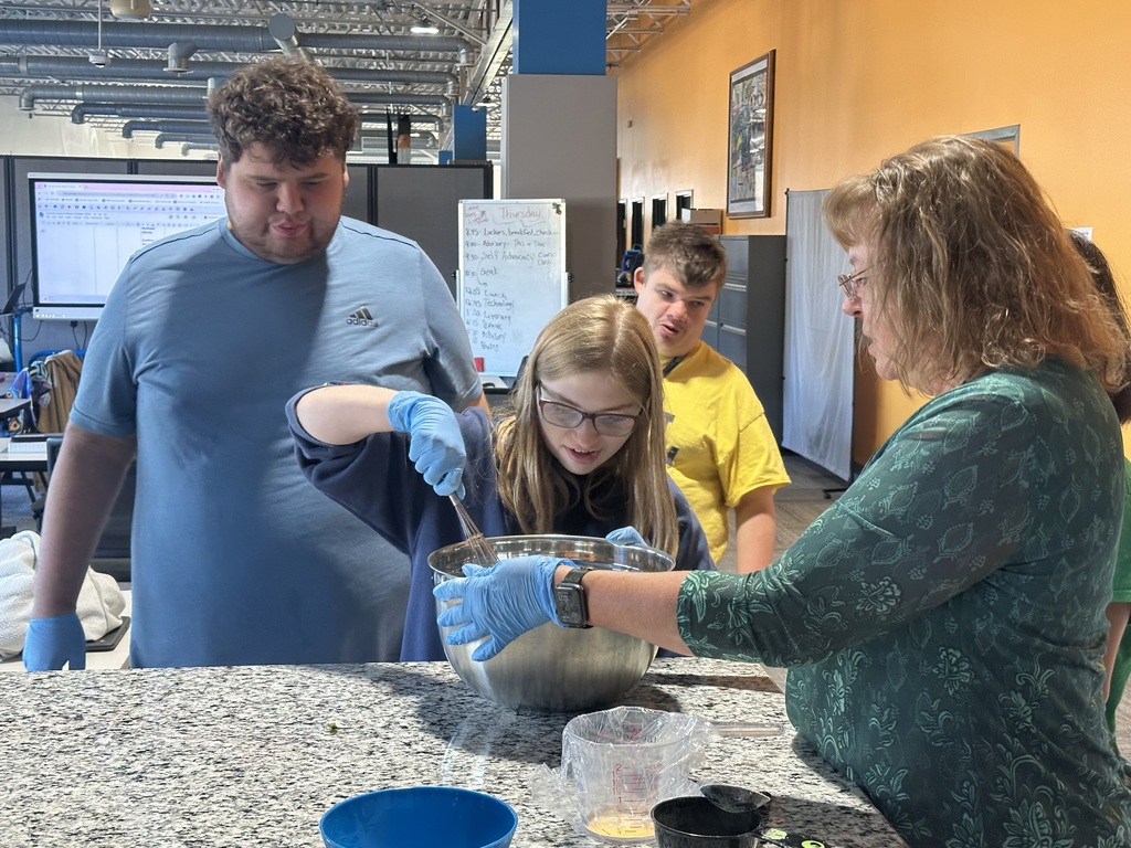 A young woman in blue gloves whisks ingredients in a metal bowl with guidance from an instructor as classmates watch.