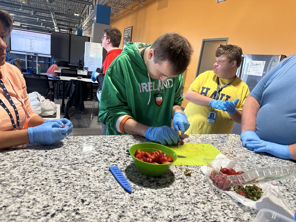 A student wearing blue gloves slices strawberries on a cutting board alongside classmates and staff at a kitchen island.
