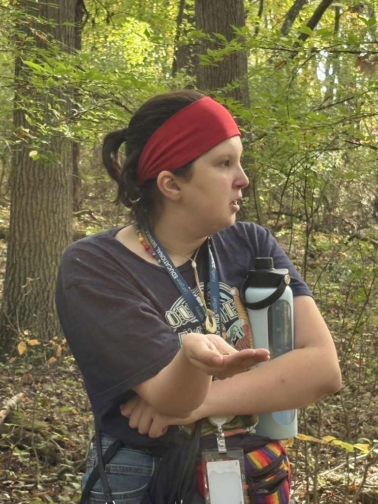 A young woman in a red headband and lanyard standing in the woods with her hand outstretched while holding a water bottle.
