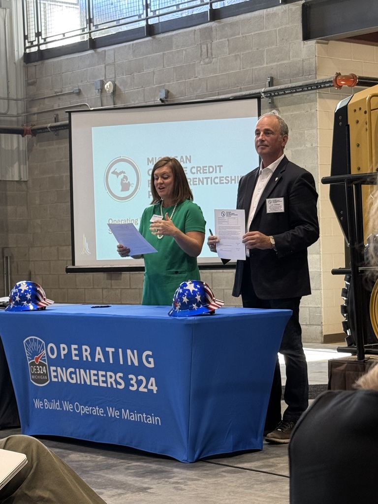 A signing ceremony at Operating Engineers Local 324 in Howell, Michigan, celebrating the Michigan College Credit for Apprenticeships Program. Two representatives stand behind a blue Operating Engineers Local 324 table holding documents, with hard hats displayed in front of them.
