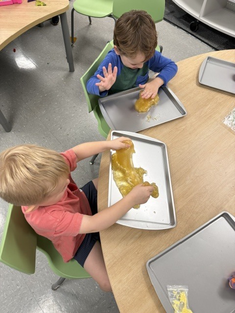 Two preschool children seated at a table stretch and explore golden sensory slime on individual metal trays.