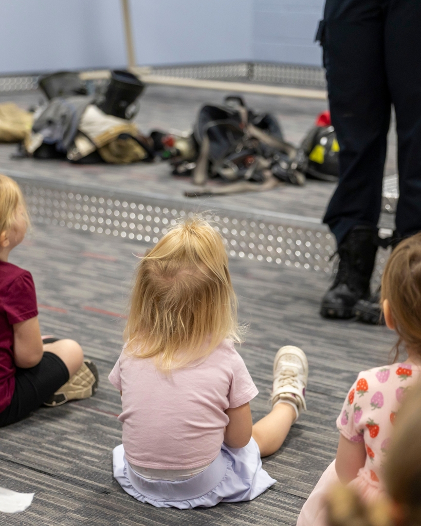 Children sit on the floor watching a demonstration with firefighter gear placed on a stage.